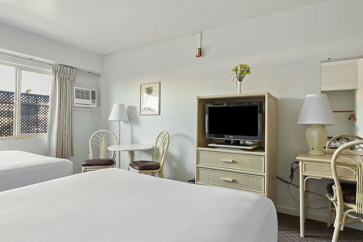 The image shows a hotel room with two beds, a TV on a cabinet, chairs around a table, a desk with a lamp, and a window with curtains.