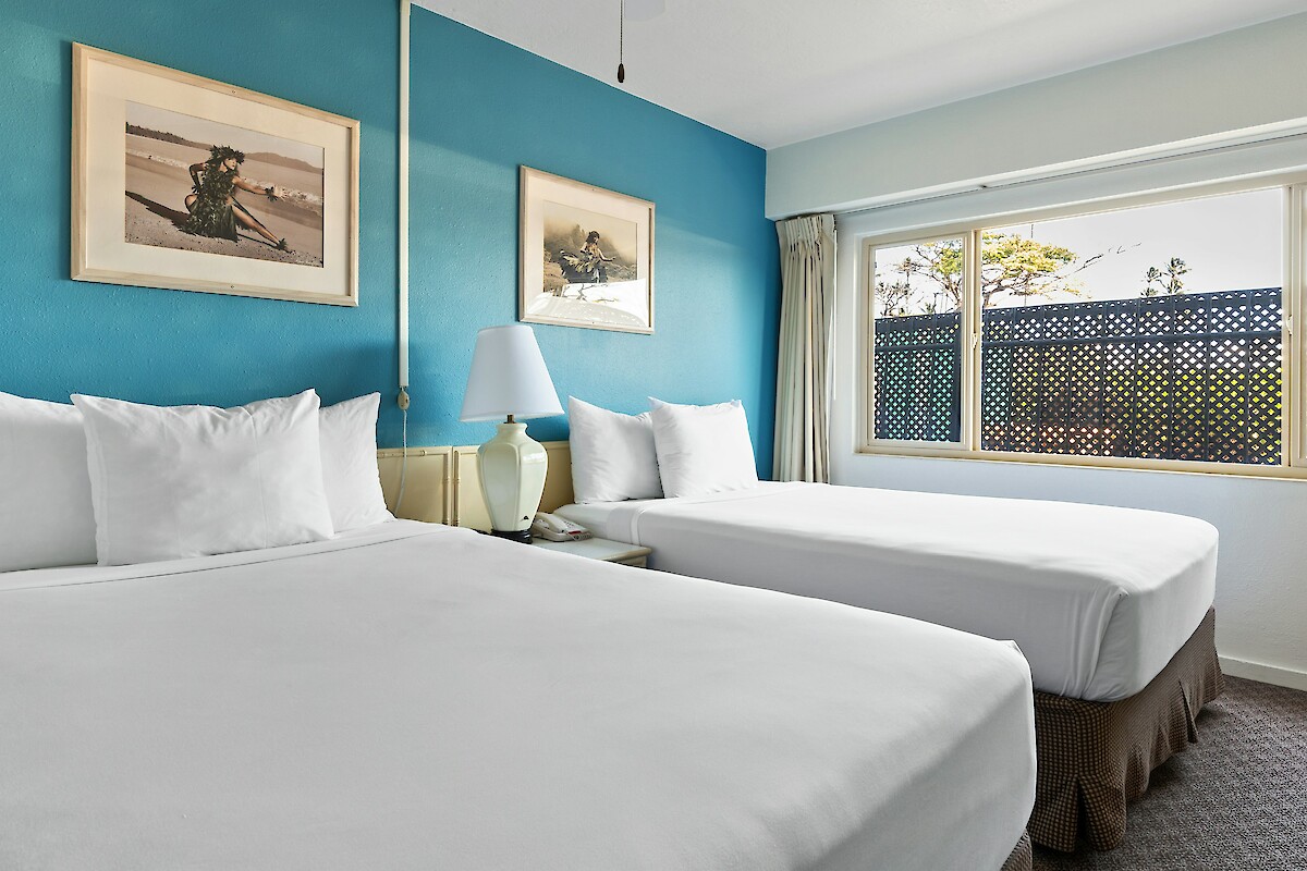 The image shows a hotel room with two neatly made beds, a blue accent wall, a lamp, framed artwork, and a window with a view outside.