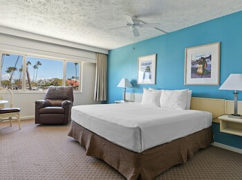 A hotel room with a bed, blue wall, two framed pictures, a ceiling fan, and a window with a view of palm trees.