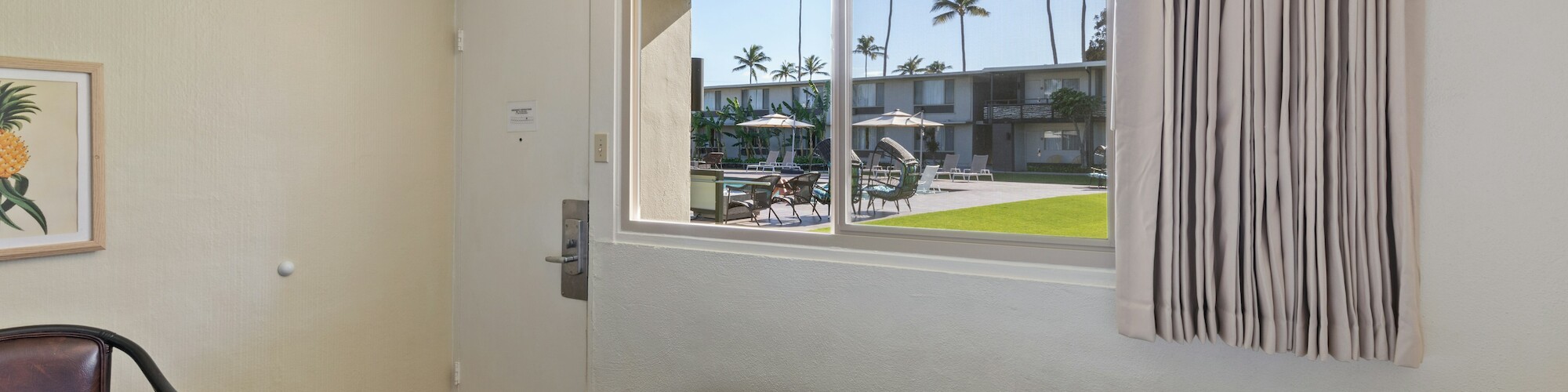 The image shows a simple room with a bed, a chair, and a window with a view of a courtyard with palm trees and outdoor seating.