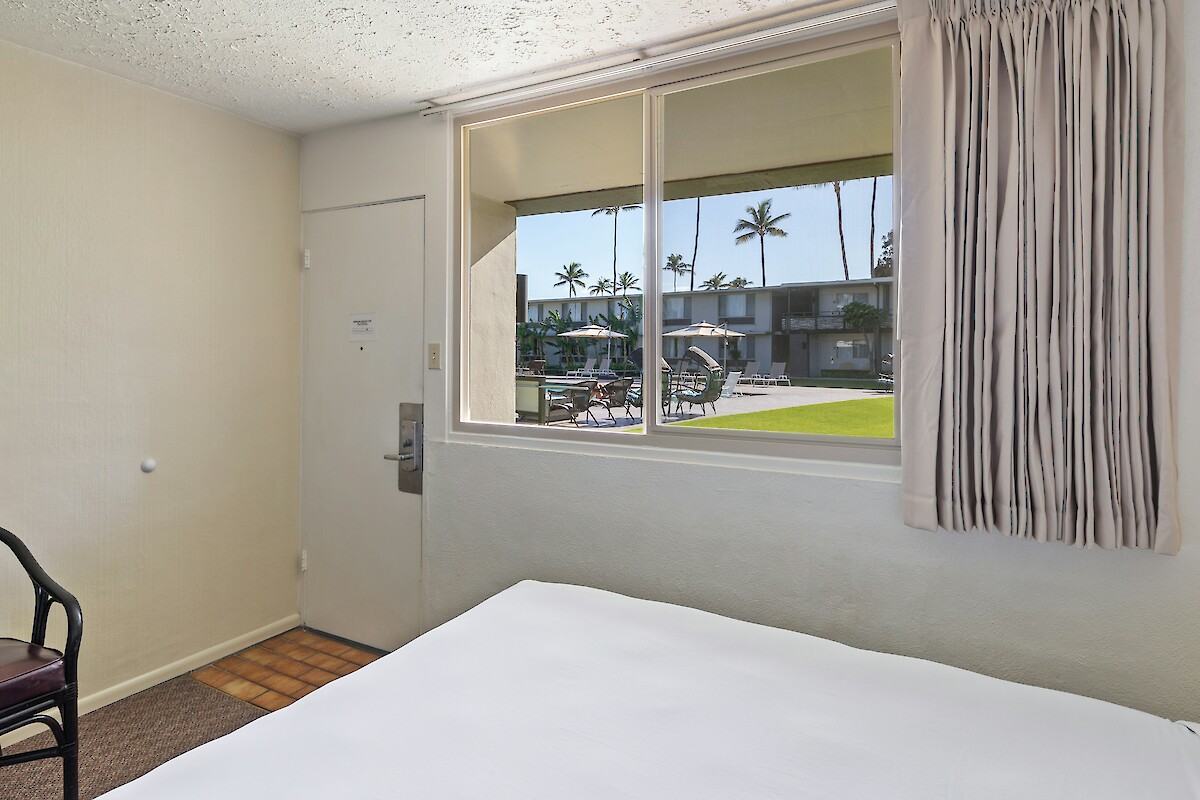 The image shows a simple hotel room with a bed, a chair, and a window view of a pool area with palm trees outside.