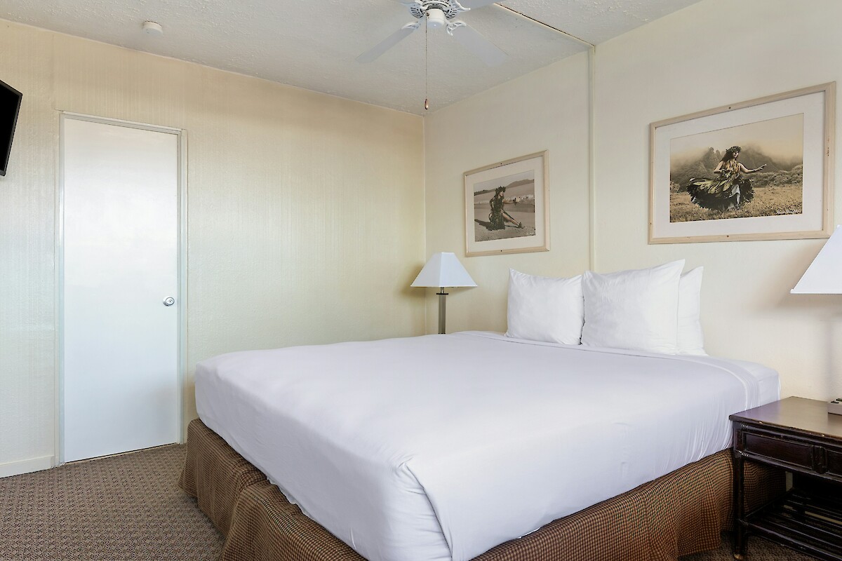 The image shows a simple bedroom with a bed, two bedside tables with lamps, framed art on the walls, and a wall-mounted TV.