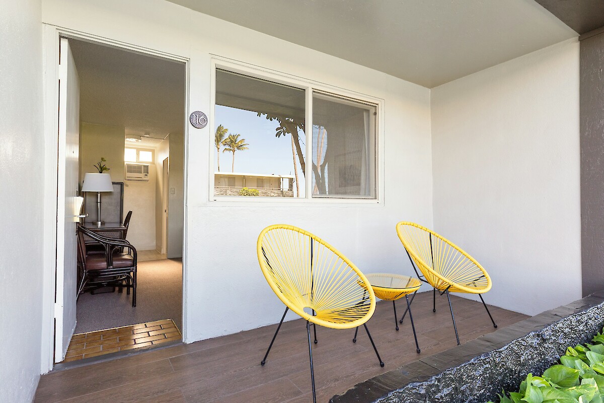A small patio with two yellow chairs, a unit number, and an open door showing an interior with furniture and lighting.