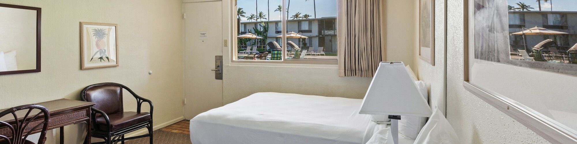 A hotel room with two beds, a table with chairs, a ceiling fan, and a window showing an outdoor scene with palm trees and umbrellas.
