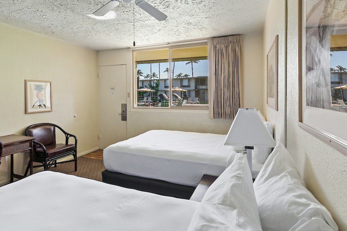 A clean hotel room with two beds, a small table and chairs. The window shows a view of palm trees outside.