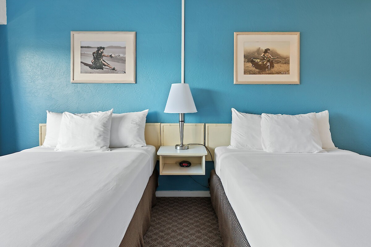 The image shows a hotel room with two neatly made beds, a shared nightstand, a lamp, and framed artwork on a blue accent wall.