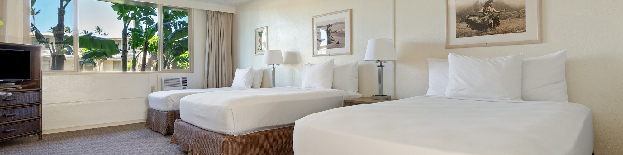 The image shows a hotel room with two large beds, a TV, a ceiling fan, and windows with a view of trees.