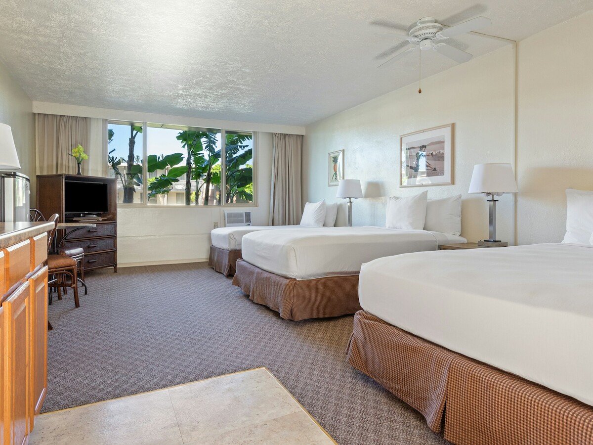 The image shows a hotel room with two large beds, a TV, wooden cabinets, and a window with a view of greenery.
