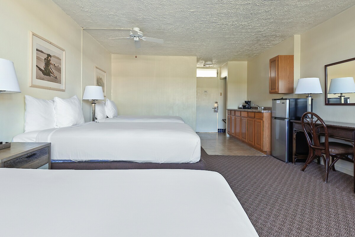 The image shows a hotel room with two beds, a small dining area, kitchenette, and a large mirror on the wall.