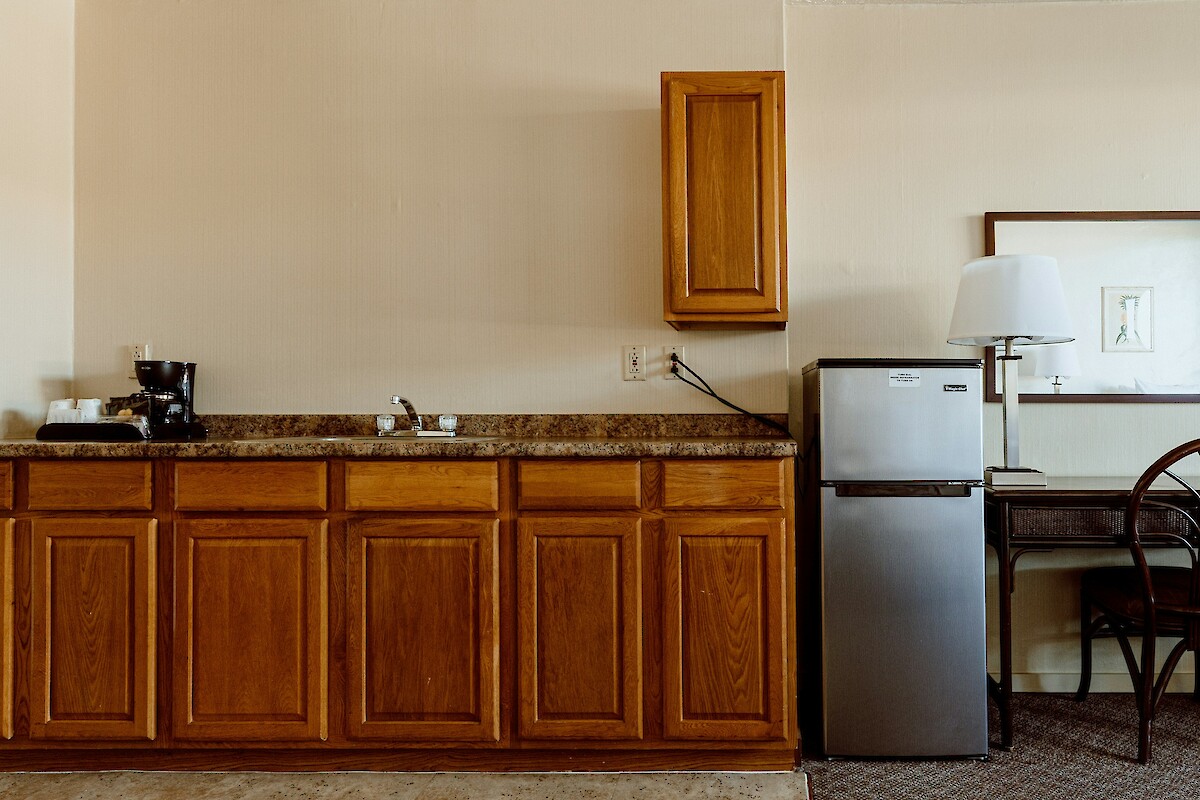 The image shows a kitchenette with wooden cabinets, a coffee maker, a sink, a small fridge, and a desk with a chair and lamp.