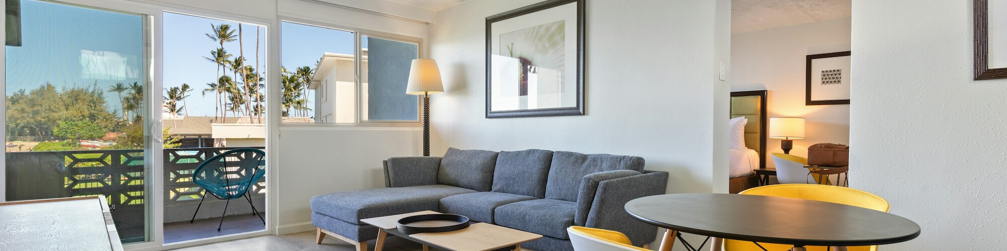 A modern living room features a gray sofa, a round table with yellow chairs, and a balcony with a lush view outside.
