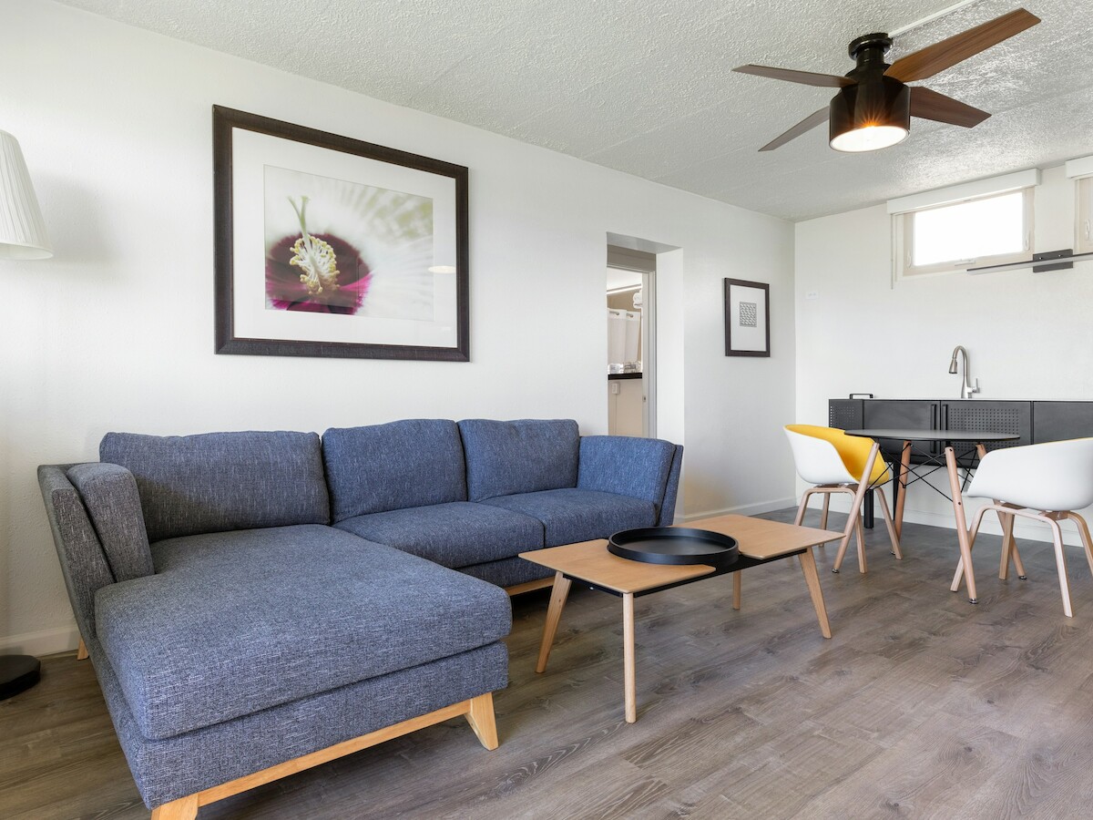 A modern living room with a blue sofa, wooden table, wall art, and dining area with chairs.