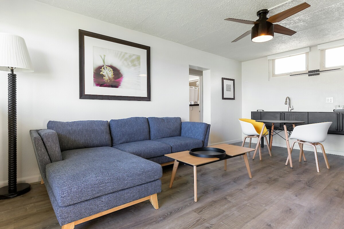 A modern living room with a blue sectional sofa, wooden table, wall art, and dining area with chairs and a minimalist kitchenette.