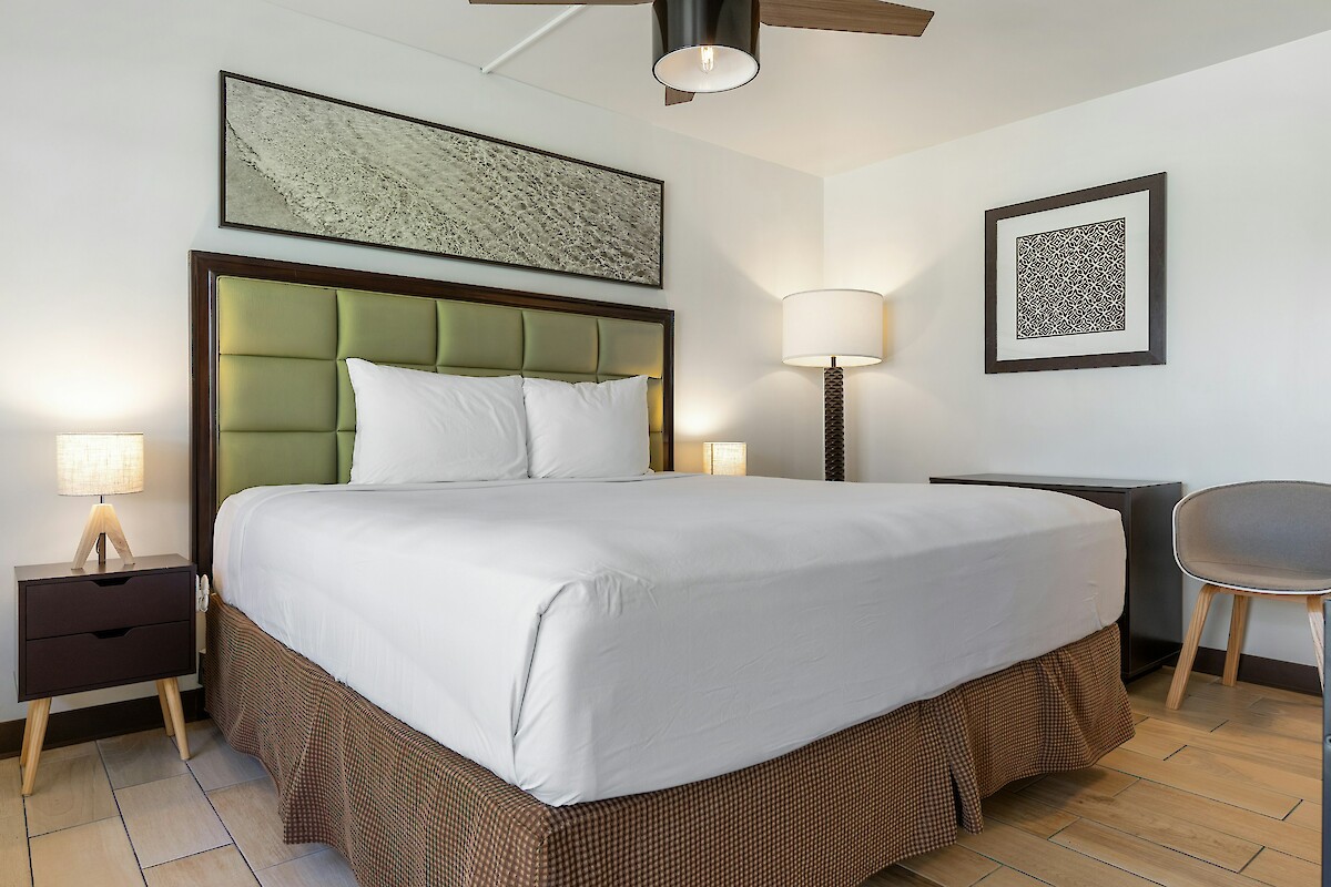 A modern hotel room with a neatly made bed, nightstands with lamps, a chair, a desk, and wall art, all in neutral tones.