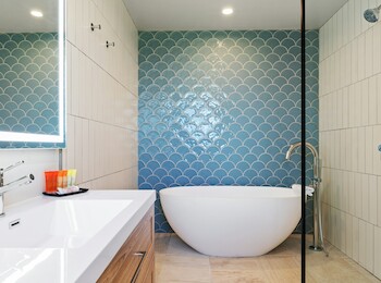 A modern bathroom features a white freestanding tub with blue fish-scale tiles, a wood vanity, a lighted mirror, and a glass shower.