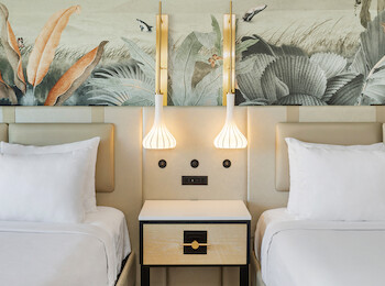 The image shows a hotel room with two beds, a decorative mural, pendant lights, and a nightstand between the beds.