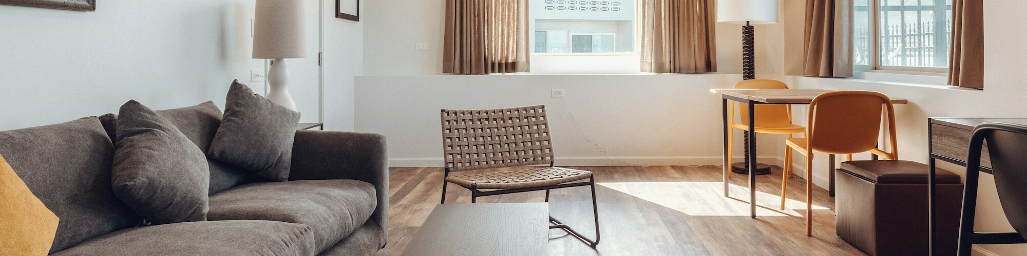 A modern living room features a gray sofa, a coffee table, a chair, a desk with a chair, and large windows with brown curtains.