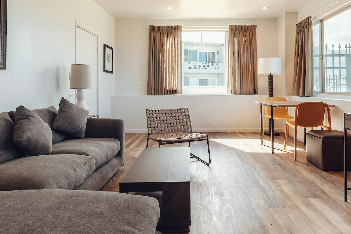 A modern living room with a gray sofa, chair, two lamps, a table, and windows with brown curtains letting in natural light.