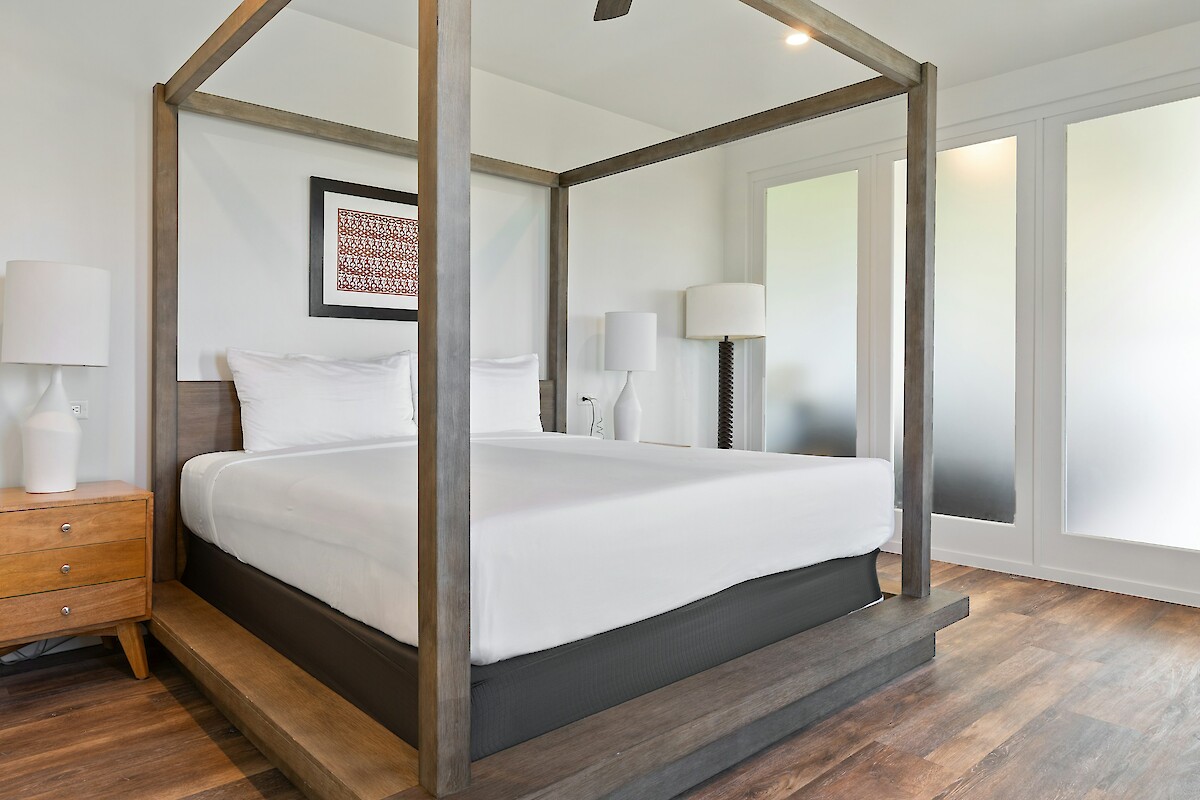The image shows a modern bedroom with a canopy bed, two white lamps, a wooden nightstand, and frosted glass doors in the background.