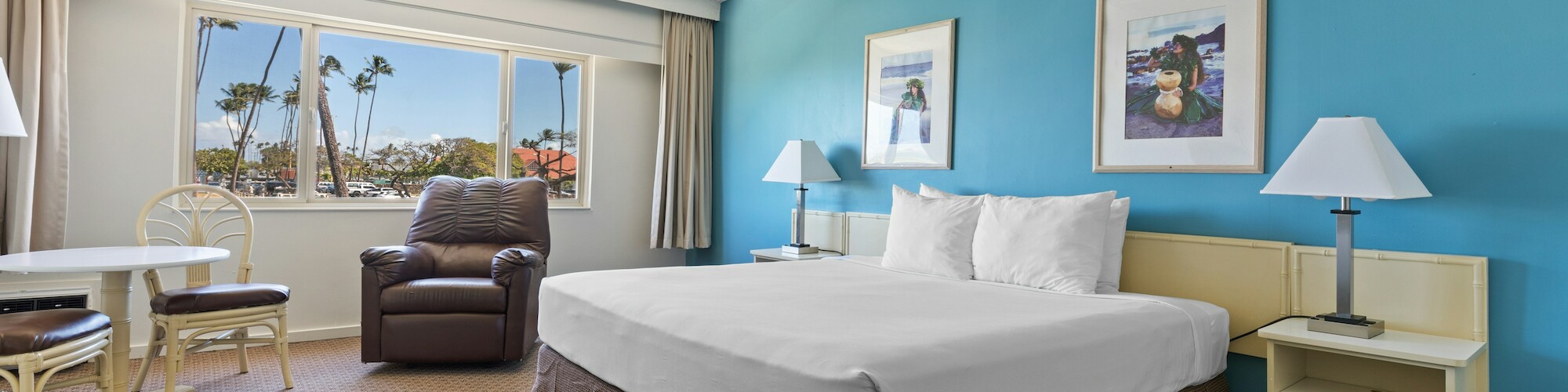 A hotel room with a large bed, turquoise wall, two lamps, artworks, a window view, a small table with chairs, and a recliner.