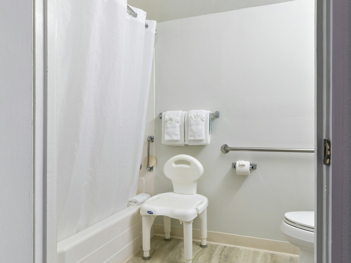 A bathroom with a shower, white curtain, towels, a toilet, a grab bar, and a shower chair beside the bathtub.