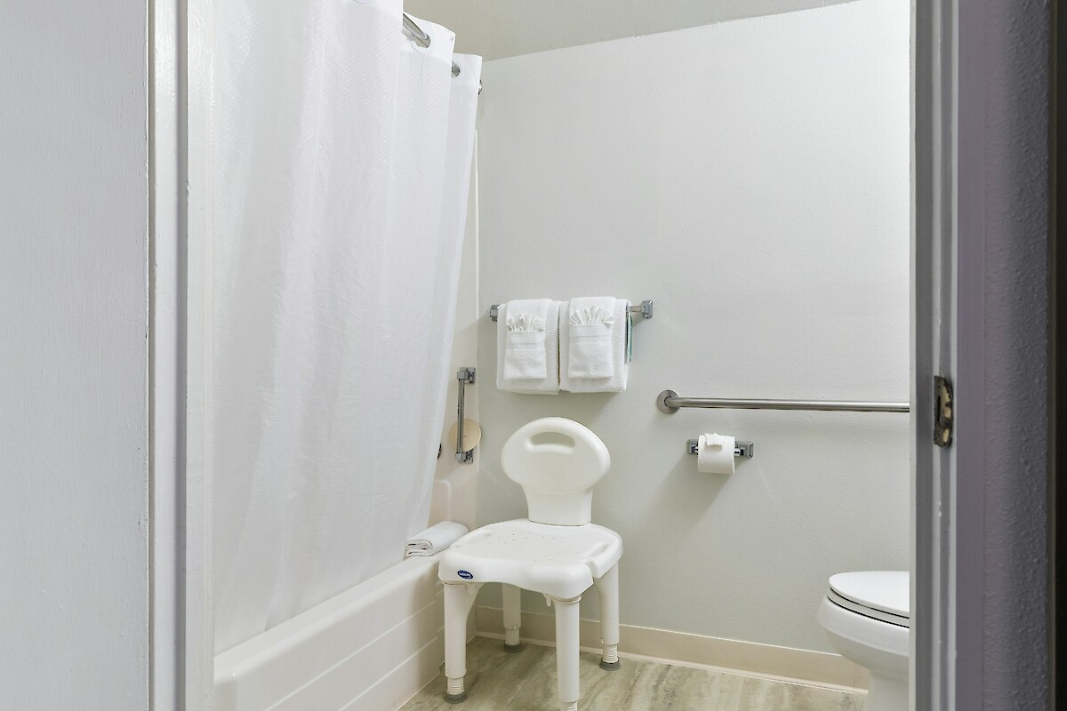 The image shows a bathroom with a shower, a white curtain, a toilet, a shower chair, towel racks with towels, and a grab bar on the wall.