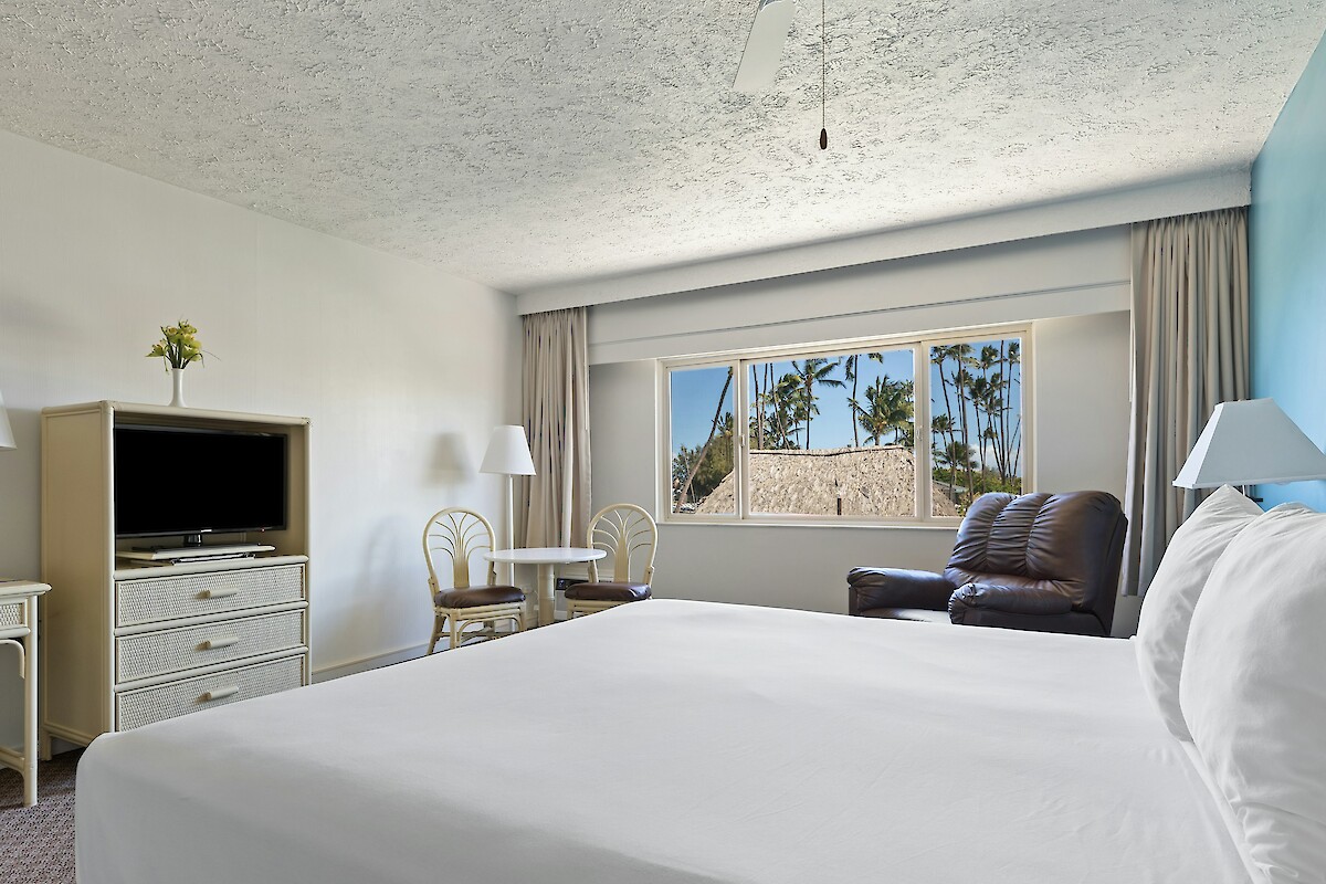 A cozy hotel room with a bed, TV, armchair, small table, chairs, and a window showing trees.