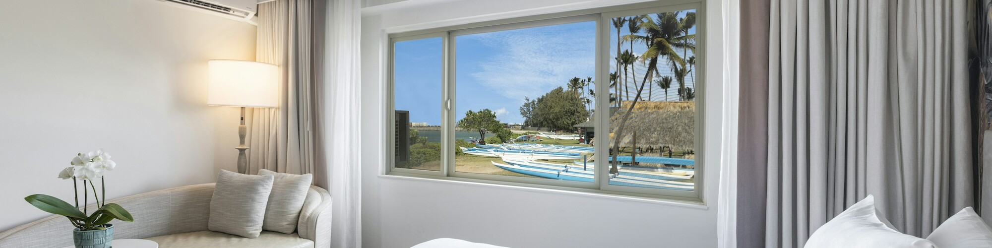 A bright, modern hotel room with a large bed, a cozy seating area, and a window offering a scenic view of a beach with palm trees.
