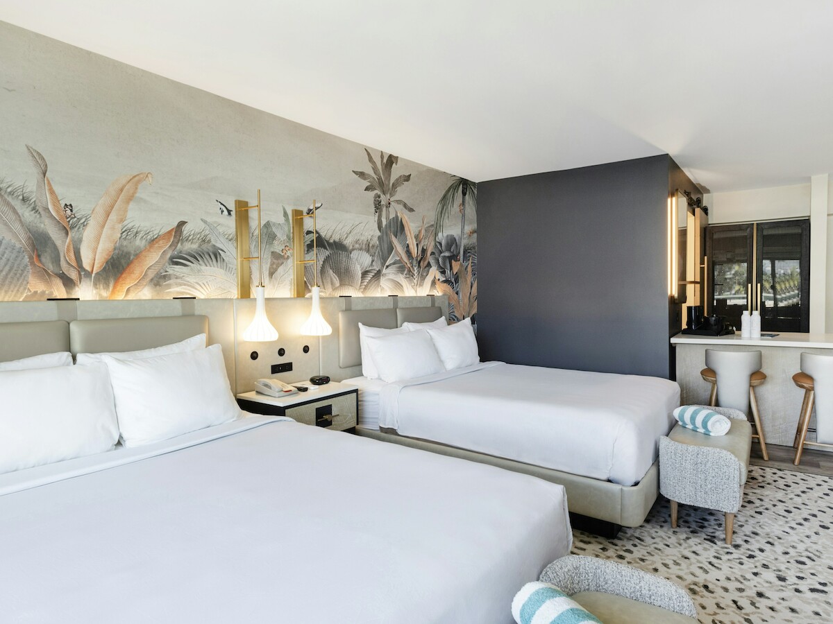 The image shows a modern hotel room with two beds, a mural on the wall, armchairs, and a kitchenette area with tall stools.