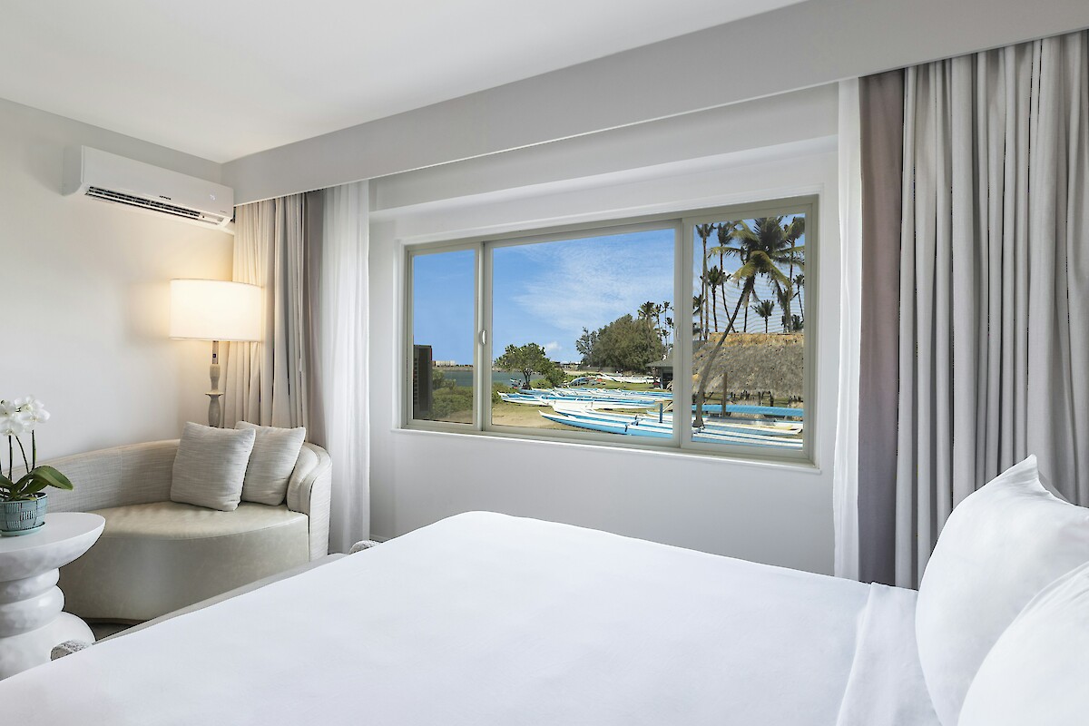 A bright hotel room with a bed, chair, and lamp, overlooking a beach scene with palm trees through the window.