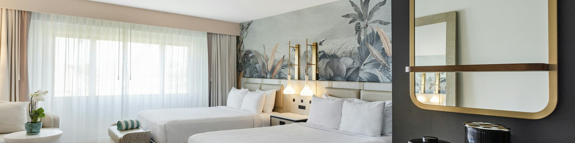 A hotel room with two beds, a wall mural, a mirror, a coffee station, and a window with sheer curtains.