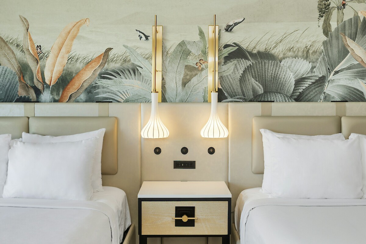 A hotel room with two beds, decorative wallpaper, modern pendant lights, and a central nightstand is shown.