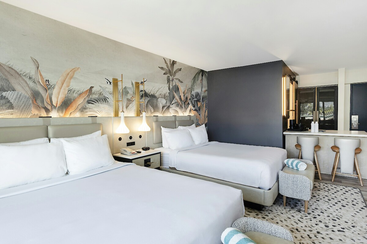 The image shows a modern hotel room with two beds, artistic wall decor, a desk area, and stylish lighting fixtures.