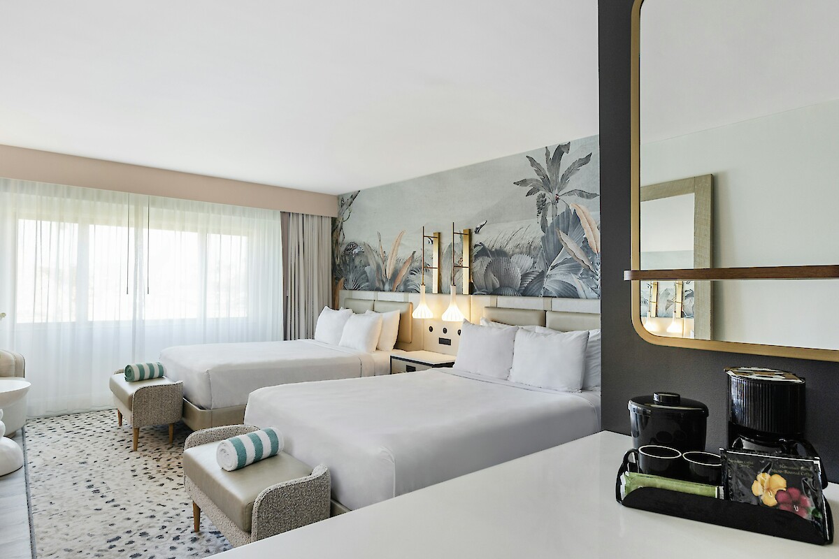 A hotel room with two beds, a large window, decorative wall art, a table with a plant, and a coffee station with mugs under a mirror.
