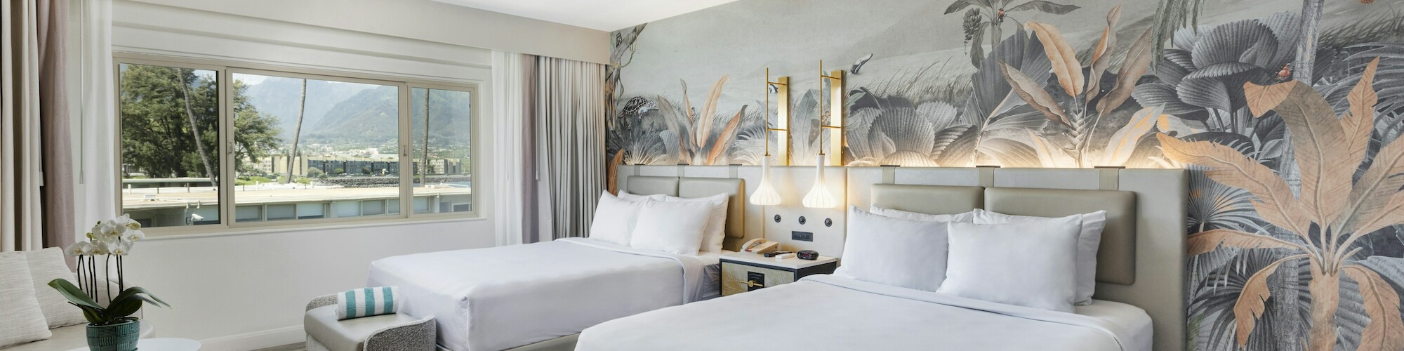 A hotel room with two beds, tropical wallpaper, a large window, and a small seating area with a table and plant on a patterned rug.