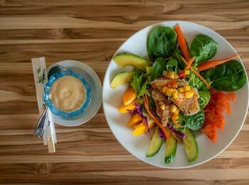 A colorful salad with greens, cucumber, tomato, corn, and dressing beside chopsticks on a wooden table.
