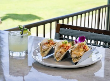 A plate with three fish tacos, garnished with a purple flower, and a cocktail with a lime wedge on a table.