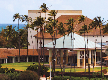 A building with a modern design surrounded by tall palm trees and a view of the ocean in the background.